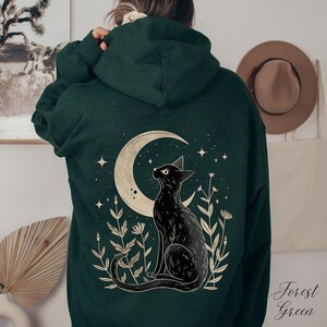 May include: A forest green hoodie with a black cat sitting under a crescent moon with stars and flowers. The text "Forest Green" is in the bottom right corner of the image.
