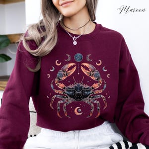 May include: Maroon-colored sweatshirt featuring a detailed crab illustration with celestial elements. The design includes a crab with intricate details, surrounded by crescent moons and a central circular design. The word "Maroon" is written in cursive on the top right.
