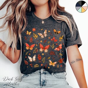 May include: A dark grey heather t-shirt with a print of colorful butterflies and leaves. The shirt has a crew neck and short sleeves.