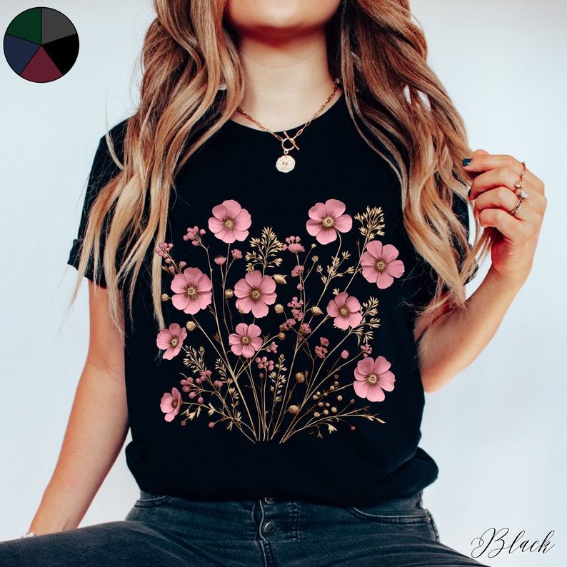 Pu&ograve; includere: Una maglietta nera con un design floreale con fiori rosa e steli dorati. I fiori sono disposti a forma di bouquet.