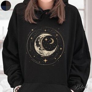 Op de afbeelding: Zwarte hoodie met een gouden halve maan en sterrenprint. De maan is omringd door een cirkel van sterren.