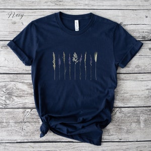 May include: A navy blue t-shirt featuring a graphic of dried flowers and grasses. The shirt has a crew neck and short sleeves.