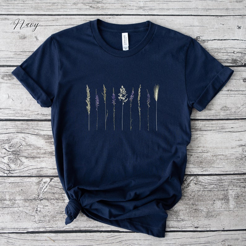 May include: A navy blue t-shirt featuring a graphic of dried flowers and grasses. The shirt has a crew neck and short sleeves.