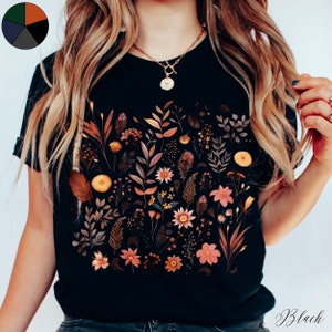 K&ouml;nnte beinhalten: Schwarzes T-Shirt mit einem floralen Druck, der verschiedene Blumen und Bl&auml;tter in Orange-, Braun- und Rosat&ouml;nen zeigt.