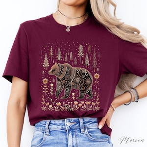 May include: Maroon t-shirt featuring a detailed bear illustration. The bear is brown with intricate patterns, set against a backdrop of gold trees and floral elements. The shirt's color is a deep maroon, with the word "Maroon" in script at the bottom.