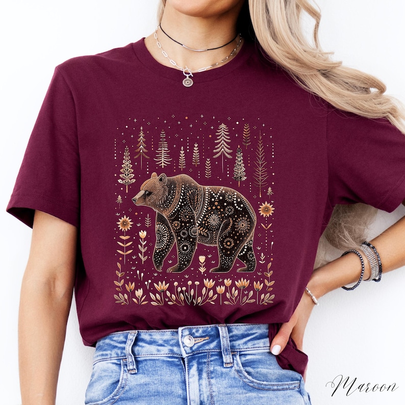 May include: Maroon t-shirt featuring a detailed bear illustration. The bear is brown with intricate patterns, set against a backdrop of gold trees and floral elements. The shirt's color is a deep maroon, with the word "Maroon" in script at the bottom.