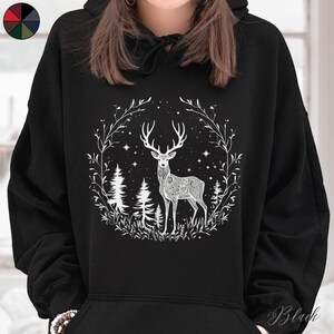 May include: Black hoodie featuring a white graphic of a deer with intricate patterns, surrounded by a wreath of leaves, stars, and trees. The design is centered on the front of the hooded sweatshirt. The hoodie has a drawstring and a kangaroo pocket.