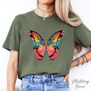 Pu&ograve; includere: T-shirt verde oliva con un vivace disegno a farfalla. La farfalla presenta un motivo colorato con sfumature di rosa, arancione, giallo e blu. Il testo "Military Green" &egrave; visibile nell'angolo in basso a destra.
