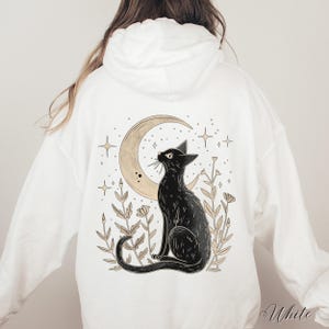 May include: White hooded sweatshirt with a black cat sitting under a crescent moon with stars and flowers. The text "White" is visible in the bottom right corner of the image.