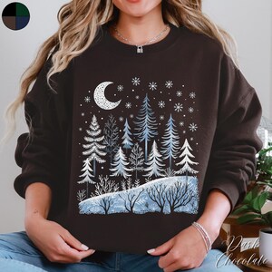 May include: A dark brown sweatshirt with a winter scene design. The design features a crescent moon, snowflakes, and a forest of blue and white trees.