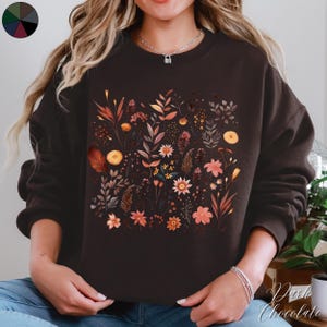 May include: A brown sweatshirt with a floral print featuring various shades of orange, yellow, and pink. The flowers are arranged in a random pattern and are surrounded by green leaves.