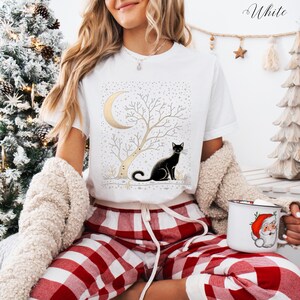 May include: White t-shirt featuring a black cat sitting under a tree with a crescent moon. The design includes a gold moon and tree, with a starry background. The word "White" is written in cursive in the upper right corner.