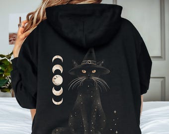 Puede incluir: Sudadera con capucha negra con un gato con sombrero de bruja y un diseño de fases lunares. El gato está adornado con estrellas. Una sudadera con capucha informal y cómoda.