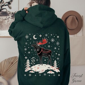 May include: Forest green hoodie featuring a winter scene with a black moose with red antlers and a decorative pattern. The design includes snowflakes, a crescent moon, and the text "Forest Green" in a script font.