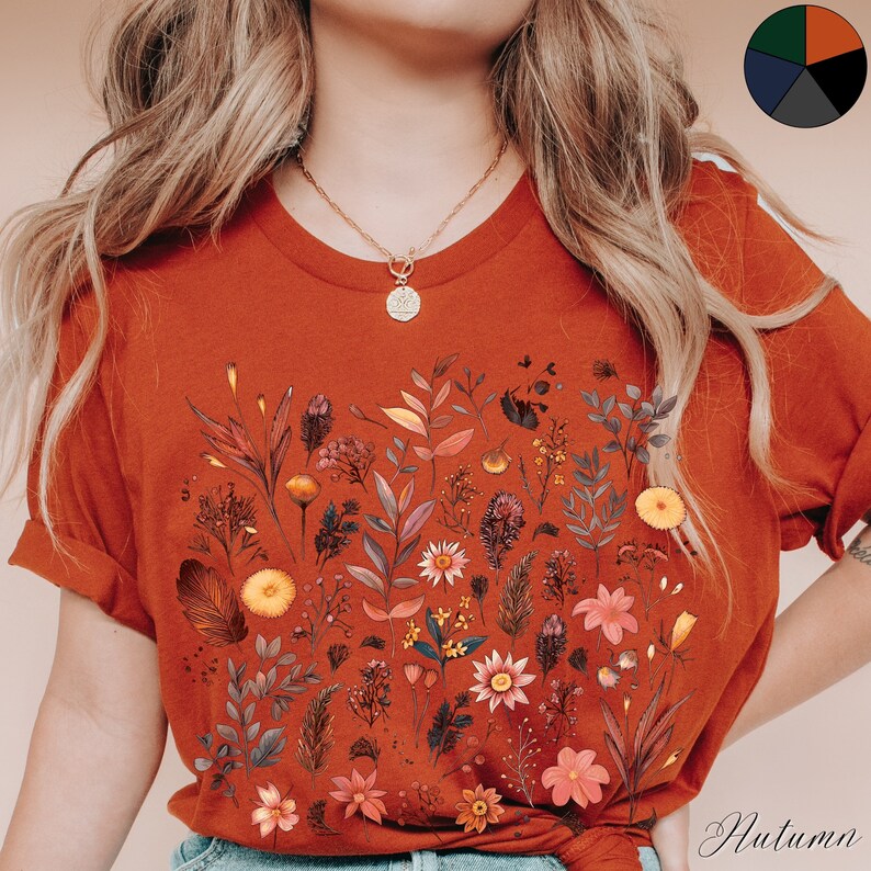 K&ouml;nnte beinhalten: Ein rostfarbenes T-Shirt mit einem Blumendruck, der verschiedene Bl&auml;tter, Bl&uuml;ten und Stiele in Braun-, Gelb- und Rott&ouml;nen zeigt. Das Shirt hat einen Rundhalsausschnitt und kurze &Auml;rmel.