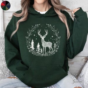 May include: Forest green hoodie featuring a white deer design within a circular frame of branches, trees, and stars. The deer has intricate patterns. The hoodie has a front pocket and a hood. The words "Forest Green" are written in cursive.