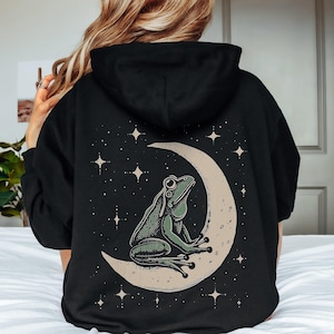 Könnte beinhalten: Schwarzer Kapuzenpullover mit einer Illustration einer grünen Frosch auf einem Mond, umgeben von Sternen. Der Frosch ist detailliert schattiert. Der Mond ist hellbeige und die Sterne sind weiß. Der Hoodie hat eine Kapuze und eine entspannte Passform.