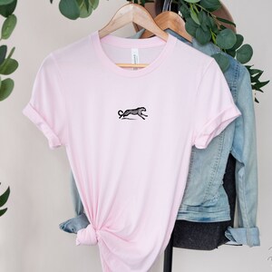 May include: A pink t-shirt with a black and white illustration of a cheetah running on the front. The shirt is on a hanger with a denim jacket behind it.