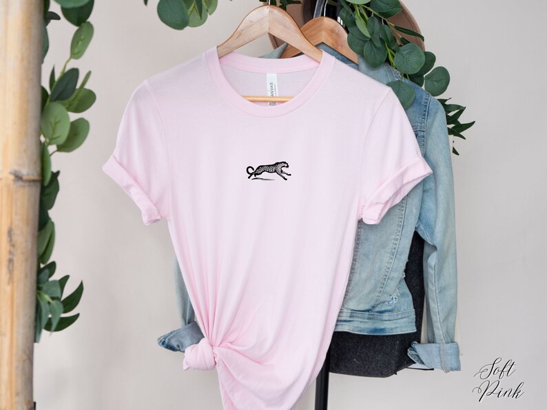 May include: A pink t-shirt with a black and white illustration of a cheetah running on the front. The shirt is on a hanger with a denim jacket behind it.