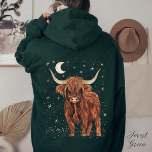 Op de afbeelding: Donkergroene hoodie met een Highland-koeienontwerp. De illustratie toont een bruine koe met lange hoorns, een maan en sterren. De tekst "Forest Green" is in script geschreven. De hoodie is donkergroen.
