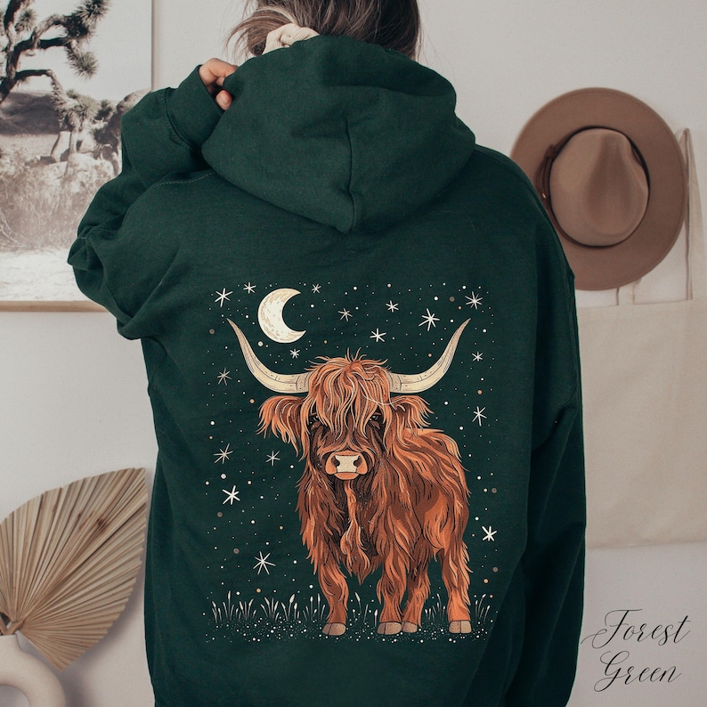 Op de afbeelding: Donkergroene hoodie met een Highland-koeienontwerp. De illustratie toont een bruine koe met lange hoorns, een maan en sterren. De tekst "Forest Green" is in script geschreven. De hoodie is donkergroen.