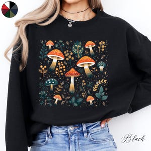 May include: Black sweatshirt with a colorful mushroom and floral print. The print features a variety of mushrooms in different colors and sizes, surrounded by leaves and flowers.