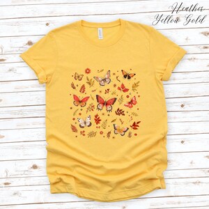May include: A yellow t-shirt with a pattern of butterflies and leaves in shades of red, orange, and brown. The text "Heather Yellow Gold" is printed in the top right corner of the image.