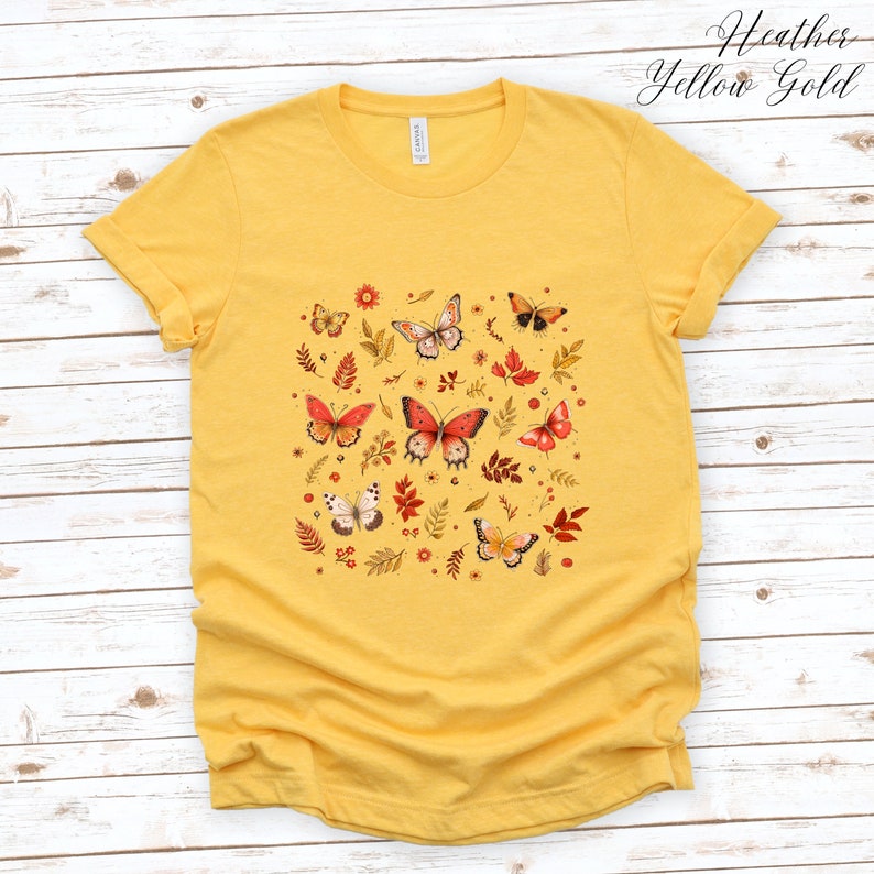 May include: A yellow t-shirt with a pattern of butterflies and leaves in shades of red, orange, and brown. The text "Heather Yellow Gold" is printed in the top right corner of the image.