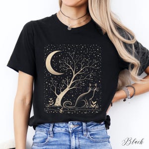 May include: Black t-shirt featuring a gold crescent moon, tree, and cat design. The illustration includes a starry night sky and the word "Black" in script at the bottom. The design is centered on the front of the shirt.