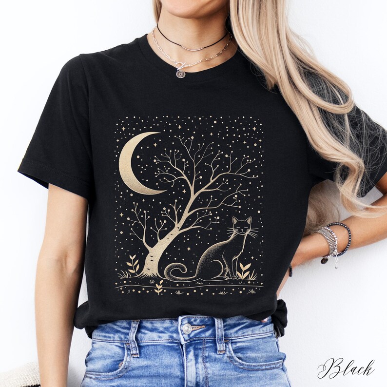 May include: Black t-shirt featuring a gold crescent moon, tree, and cat design. The illustration includes a starry night sky and the word "Black" in script at the bottom. The design is centered on the front of the shirt.