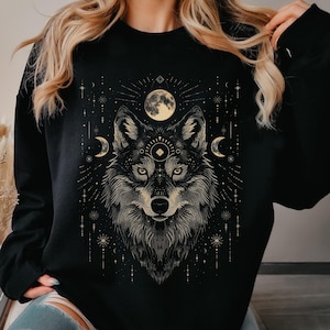 Peut inclure: Sweat-shirt noir avec une illustration détaillée d'un loup, une pleine lune, des croissants de lune et des étoiles. Le design est en beige et or, créant un thème céleste. Le sweat-shirt est de style décontracté et confortable.