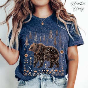 May include: Heather Navy t-shirt featuring a detailed brown bear design with floral and geometric patterns. The bear is set against a backdrop of stylized trees and flowers. The shirt has the words "Heather Navy" in the upper right corner.
