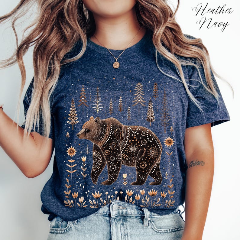 May include: Heather Navy t-shirt featuring a detailed brown bear design with floral and geometric patterns. The bear is set against a backdrop of stylized trees and flowers. The shirt has the words "Heather Navy" in the upper right corner.
