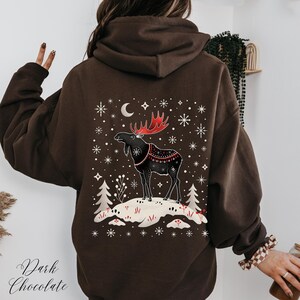 May include: Dark chocolate brown hoodie featuring a winter scene with a moose graphic. The moose has red antlers and is wearing a decorative blanket. The design includes snowflakes, a crescent moon, and the text "Dark Chocolate".
