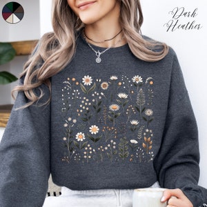 Könnte beinhalten: Ein dunkelgraues Sweatshirt mit einem Blumenmuster aus weißen Gänseblümchen und grünen Blättern. Der Text "Dark Heather" ist in der oberen rechten Ecke des Bildes sichtbar.