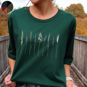 May include: A dark green t-shirt featuring a row of dried flowers and grasses printed on the front. The flowers are purple and white, and the grasses are green and brown.