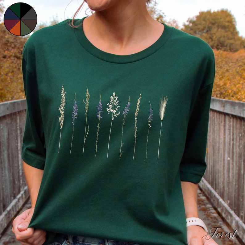 May include: A dark green t-shirt featuring a row of dried flowers and grasses printed on the front. The flowers are purple and white, and the grasses are green and brown.