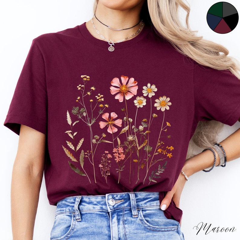 May include: A maroon t-shirt with a floral print featuring dried flowers in shades of pink, white, and brown. The flowers are arranged in a cluster on the front of the shirt.