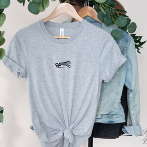 May include: A gray t-shirt with a black embroidered image of a running leopard. The shirt is hanging on a hanger with a denim jacket.