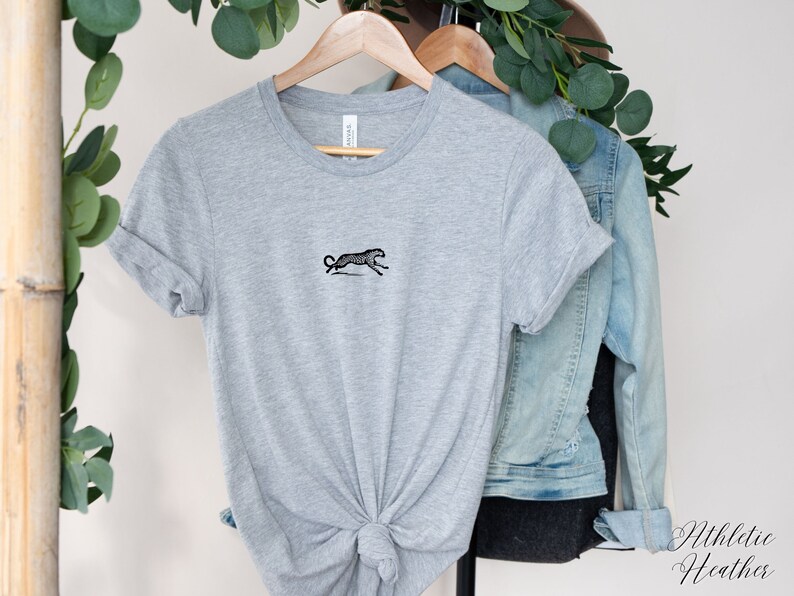 May include: A gray t-shirt with a black embroidered image of a running leopard. The shirt is hanging on a hanger with a denim jacket.