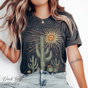 May include: Dark gray heather t-shirt featuring a desert-themed graphic. The design includes a sun with a face, cacti, and the text "Dark Greg Heather". The shirt is made of a soft material.