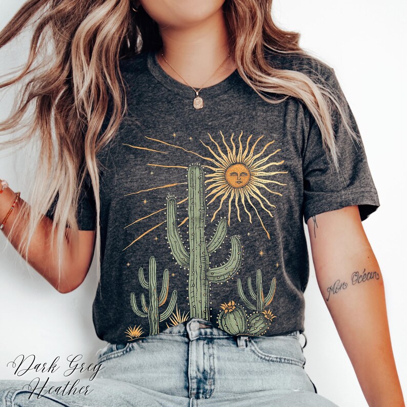 May include: Dark gray heather t-shirt featuring a desert-themed graphic. The design includes a sun with a face, cacti, and the text "Dark Greg Heather". The shirt is made of a soft material.