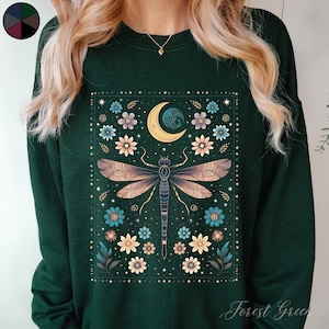 May include: A forest green sweatshirt featuring a printed design of a dragonfly, moon and flowers. The design is surrounded by a border of stars.