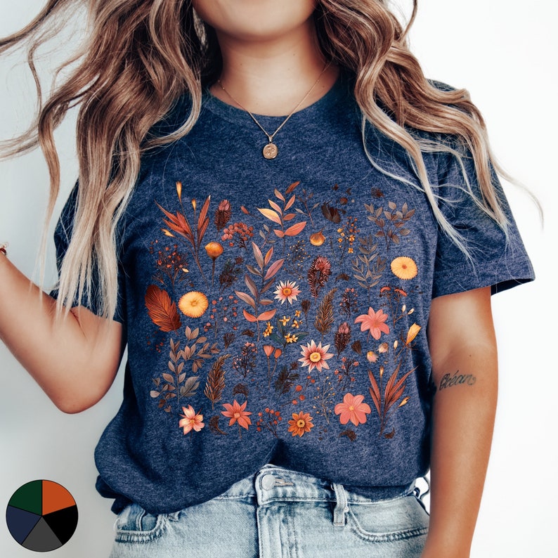 K&ouml;nnte beinhalten: Ein marineblaues T-Shirt mit einem Blumenmuster, das verschiedene Blumen und Bl&auml;tter in Orange-, Gelb- und Rosat&ouml;nen zeigt. Das Shirt hat einen Rundhalsausschnitt und kurze &Auml;rmel.