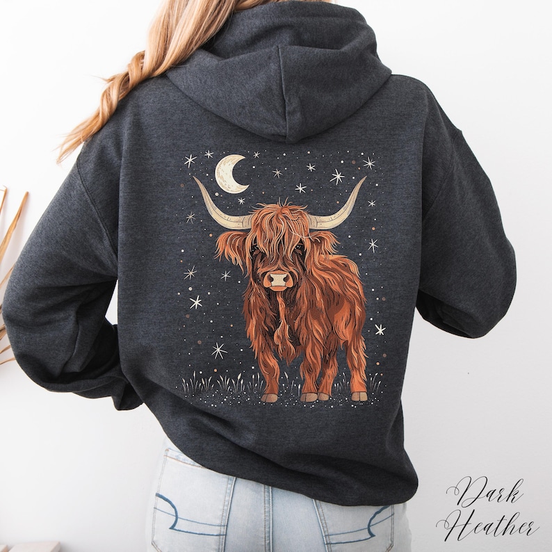 Op de afbeelding: Donkergrijze hoodie met een gedetailleerde illustratie van een Schotse Hooglandkoe, met lange hoorns, onder een maan en sterren. De koe is in tinten bruin en oranje, tegen een donkere achtergrond. De woorden "Dark Heather" zijn in script geschreven.