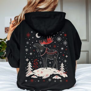 May include: Black hoodie featuring a detailed graphic of a moose with red antlers and a decorative blanket. The design includes snowflakes, a crescent moon, and stylized trees. The moose stands on a snowy hill, creating a winter theme.