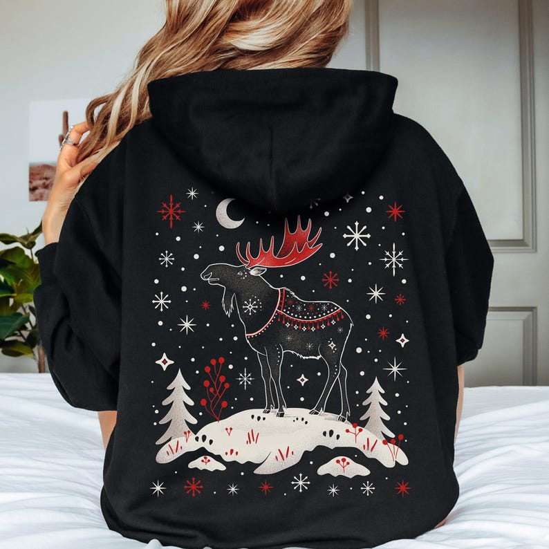 May include: Black hoodie featuring a detailed graphic of a moose with red antlers and a decorative blanket. The design includes snowflakes, a crescent moon, and stylized trees. The moose stands on a snowy hill, creating a winter theme.
