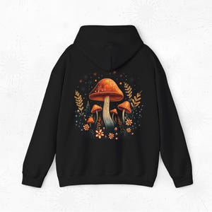 Op de afbeelding: Zwarte hoodie met een kleurrijk paddenstoelontwerp. Het ontwerp toont oranje en witte paddenstoelen, omringd door bloemen en bladeren. De hoodie heeft een capuchon en lange mouwen.