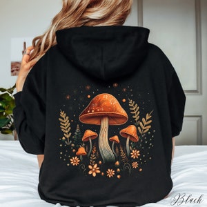 Op de afbeelding: Zwarte hoodie met een kleurrijk paddestoelontwerp op de achterkant. Het ontwerp toont oranje paddenstoelen met witte stippen, omringd door bladeren en bloemen. Het woord "Black" staat rechtsonder gedrukt.