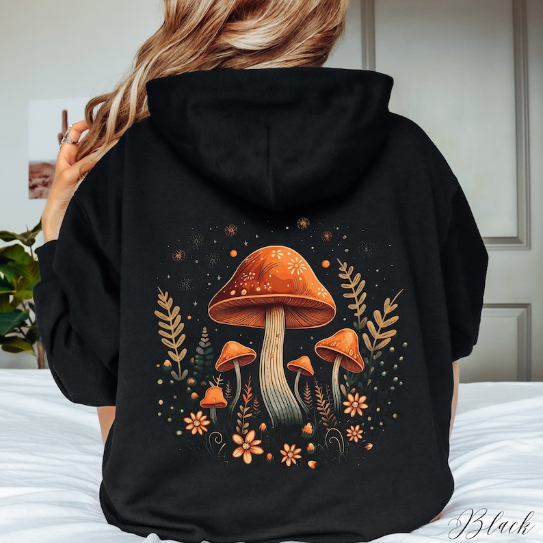 Op de afbeelding: Zwarte hoodie met een kleurrijk paddestoelontwerp op de achterkant. Het ontwerp toont oranje paddenstoelen met witte stippen, omringd door bladeren en bloemen. Het woord "Black" staat rechtsonder gedrukt.